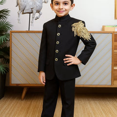 Young boy in a formal black suit with gold accents standing in a room with decorative elements.