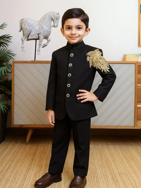 Young boy in a formal black suit with gold accents standing in a room with decorative elements.