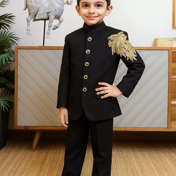 Young boy in a formal black suit with gold accents standing in a room with decorative elements.