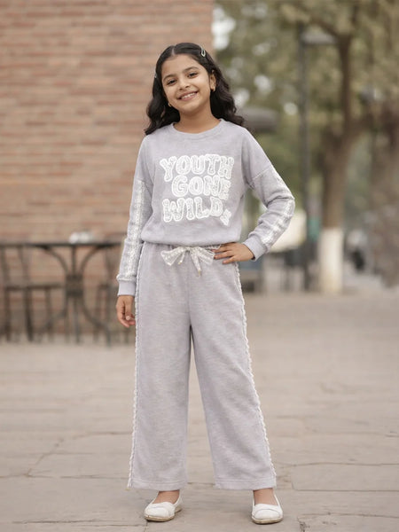 Young girl wearing a gray sweatshirt and pants with text, standing outdoors.