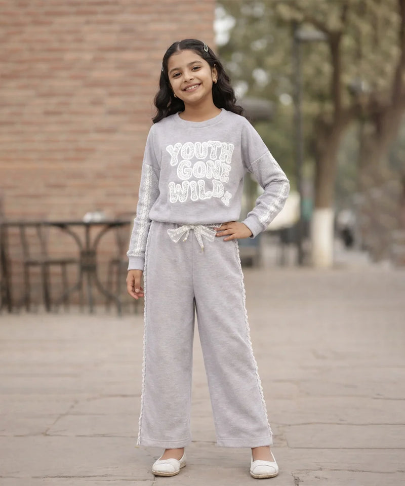 Young girl wearing a gray sweatshirt and pants with text, standing outdoors.