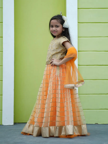 Young girl in an orange and gold traditional outfit standing against a green wall.