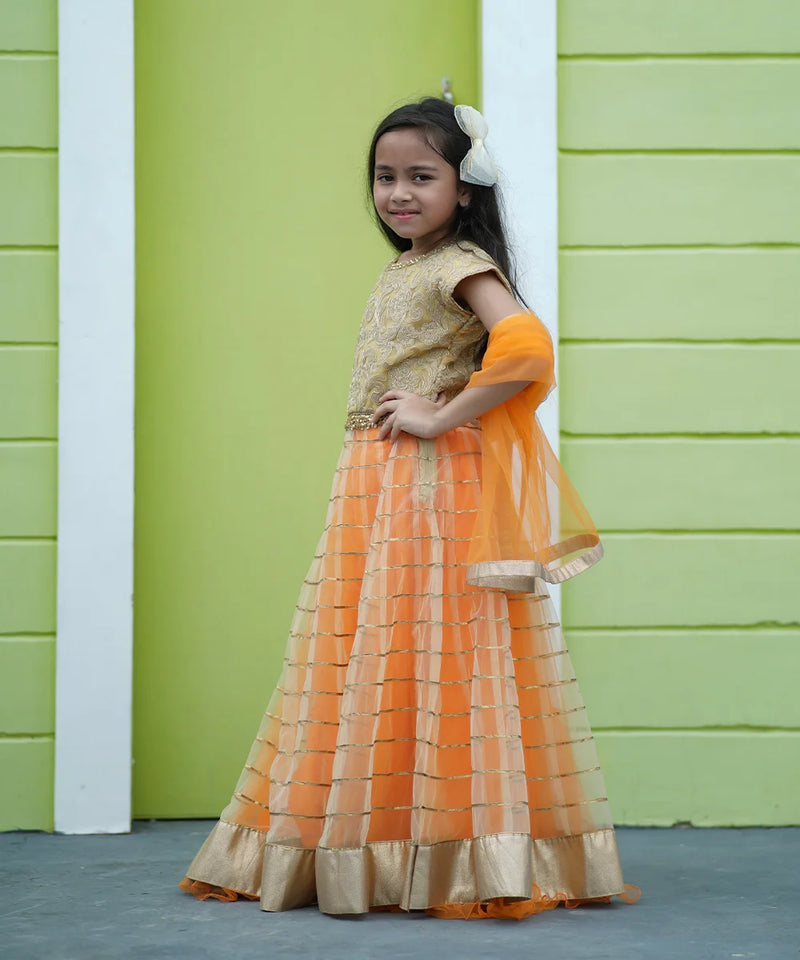 Young girl in an orange and gold traditional outfit standing against a green wall.