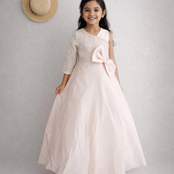 Young girl in a light pink dress with a bow, standing against a plain background.