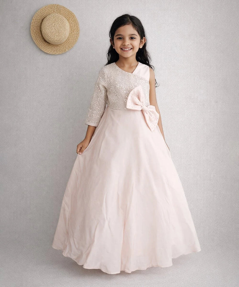 Young girl in a light pink dress with a bow, standing against a plain background.