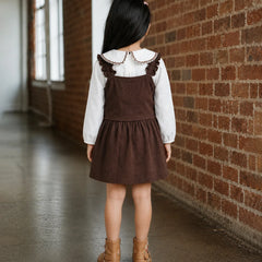 A young girl is shown from behind wearing a brown corduroy pinafore dress over a white long-sleeve shirt with a ruffled collar, paired with tan ankle boots. She is standing in a hallway with a brick wall on one side and windows on the other.