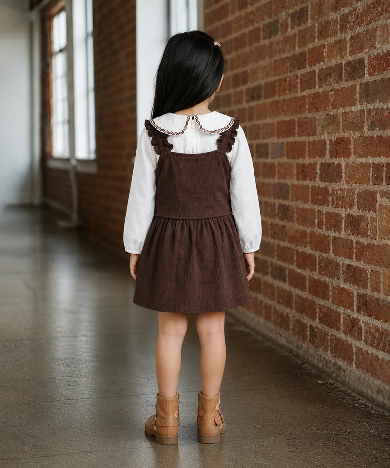 A young girl is shown from behind wearing a brown corduroy pinafore dress over a white long-sleeve shirt with a ruffled collar, paired with tan ankle boots. She is standing in a hallway with a brick wall on one side and windows on the other.