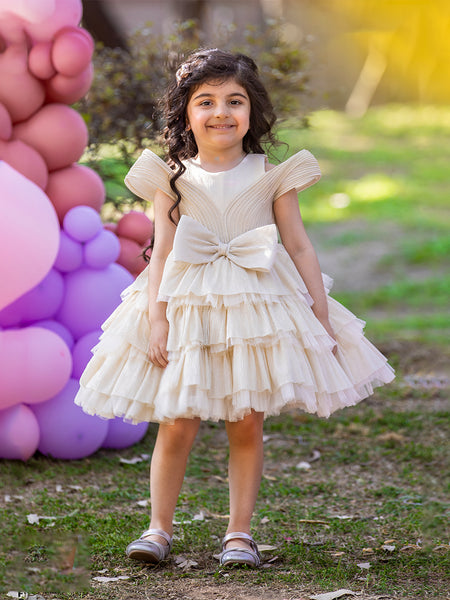 A young girl wearing a beige, white, and pink party wear birthday frock with short sleeves and a bow tie at the waist, standing in front of a backdrop with pink and purple balloons.