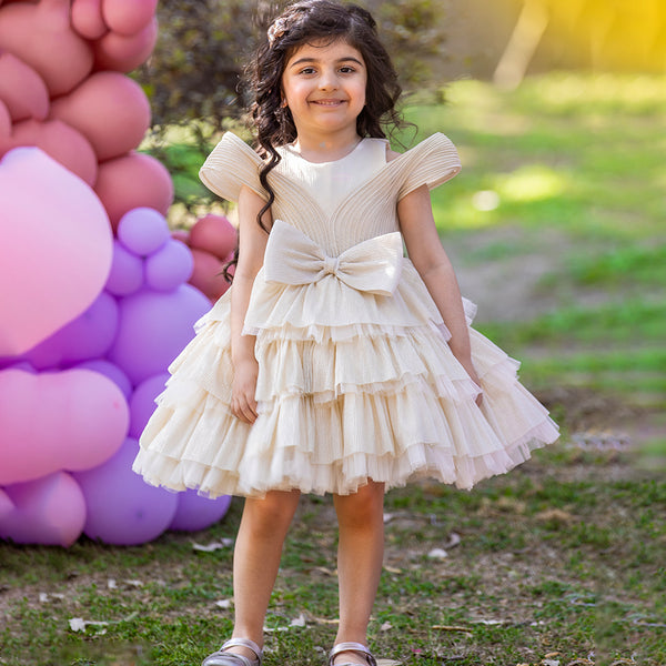 A young girl wearing a beige, white, and pink party wear birthday frock with short sleeves and a bow tie at the waist, standing in front of a backdrop with pink and purple balloons.
