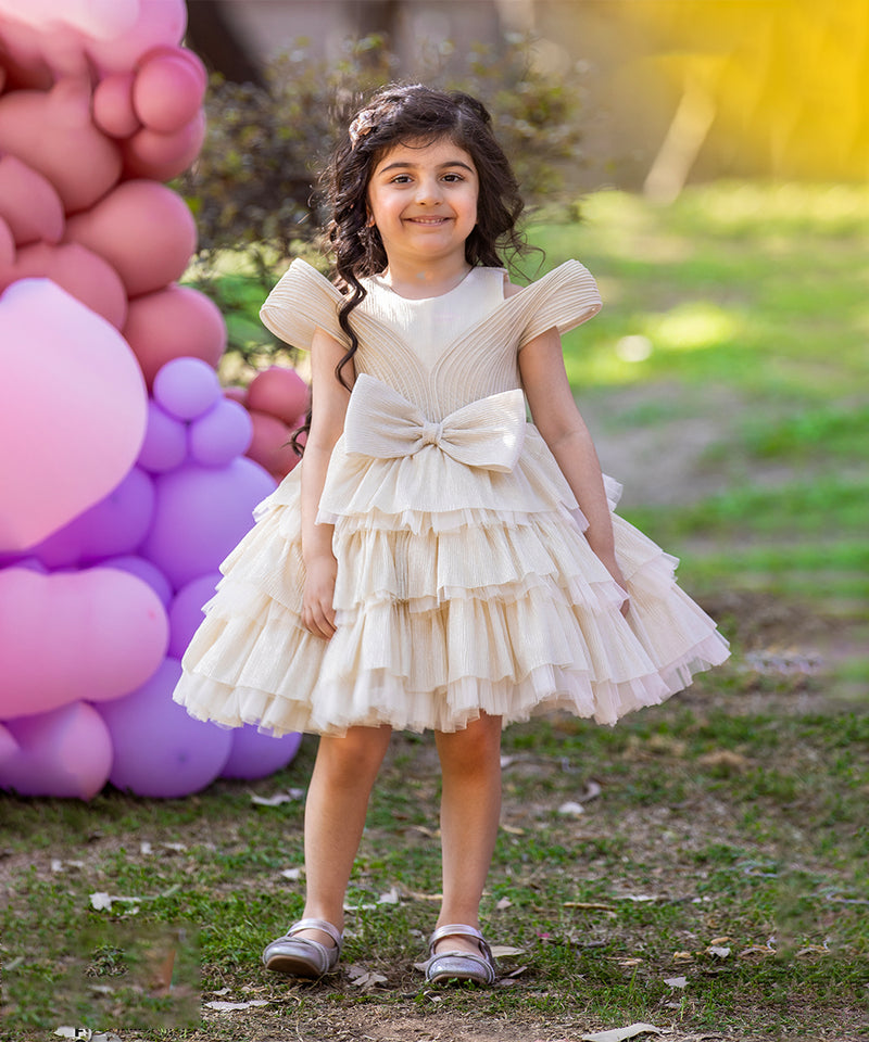 A young girl wearing a beige, white, and pink party wear birthday frock with short sleeves and a bow tie at the waist, standing in front of a backdrop with pink and purple balloons.