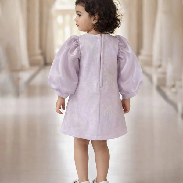 Child wearing a light purple dress with puffed sleeves in a softly lit room.