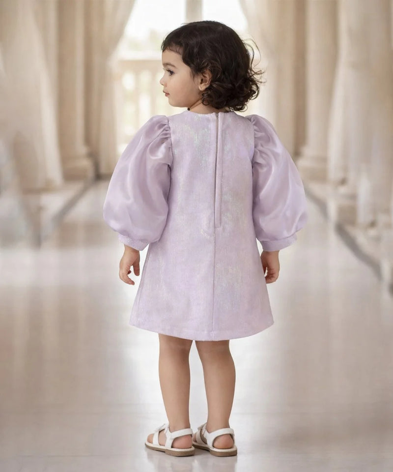 Child wearing a light purple dress with puffed sleeves in a softly lit room.