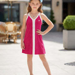 A young girl wearing a bright pink sleeveless tennis dress with white trim and a matching light blue headband, standing outdoors in a sunny, modern plaza setting.
