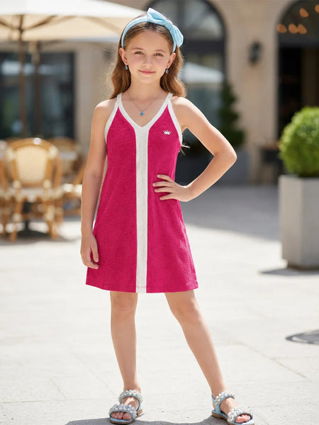 A young girl wearing a bright pink sleeveless tennis dress with white trim and a matching light blue headband, standing outdoors in a sunny, modern plaza setting.