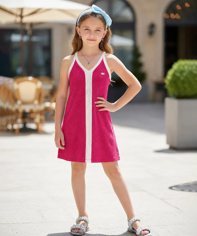 A young girl wearing a bright pink sleeveless tennis dress with white trim and a matching light blue headband, standing outdoors in a sunny, modern plaza setting.