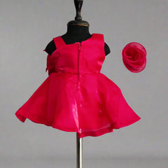 A pink colored kids' frock displayed on a mannequin, featuring a big bow on the shoulder, a flower attached with shiny lace on the waist, and a fabric belt at the back. A matching pink hair band is also shown.