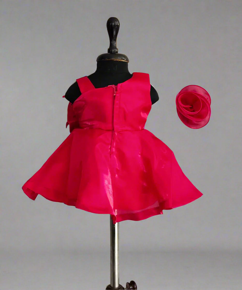 A pink colored kids' frock displayed on a mannequin, featuring a big bow on the shoulder, a flower attached with shiny lace on the waist, and a fabric belt at the back. A matching pink hair band is also shown.