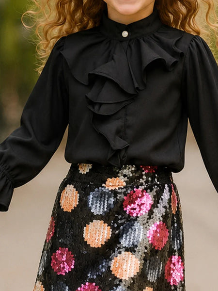 A child wearing a black long-sleeve blouse with layered ruffles and a high collar, paired with a black sequined skirt featuring large multicolored polka dots, against a blurred outdoor background.