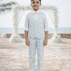 Young boy in a light blue vest and pants standing on a beach with decorative arches in the background.