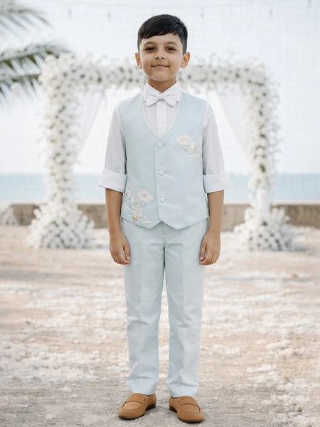 Young boy in a light blue vest and pants standing on a beach with decorative arches in the background.