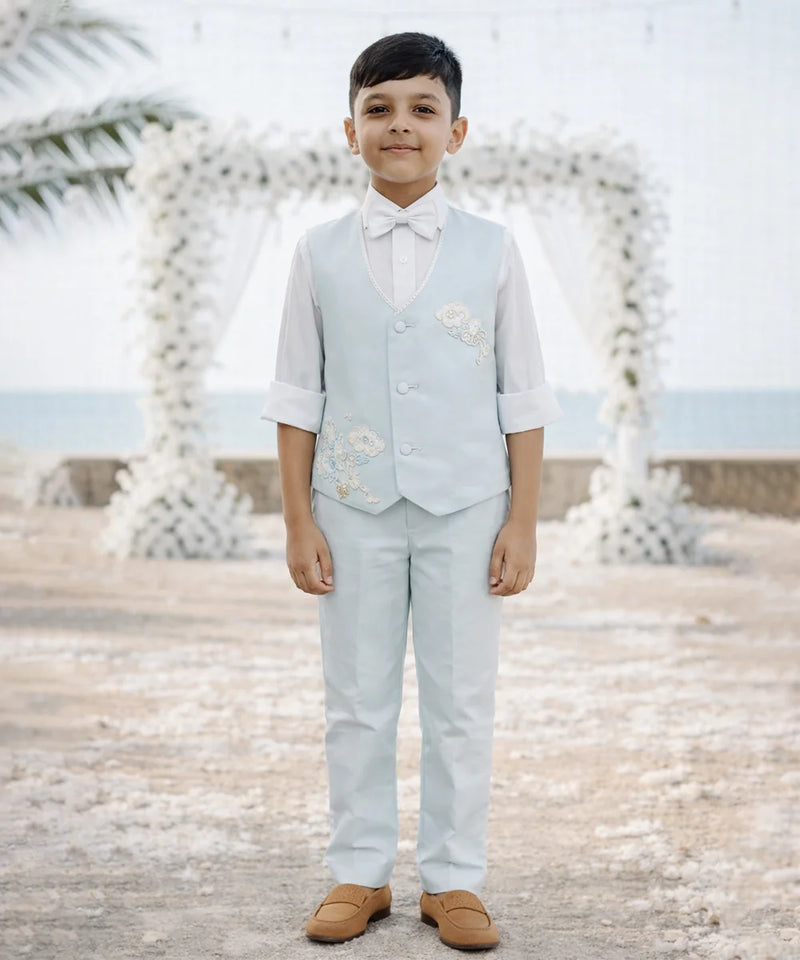 Young boy in a light blue vest and pants standing on a beach with decorative arches in the background.