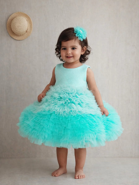 Young girl in a turquoise dress with a ruffled skirt standing against a plain background.
