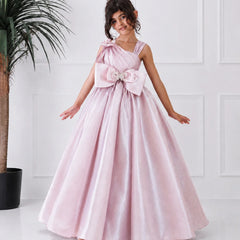 Young girl in a pink formal gown with a large bow standing against a white wall.
