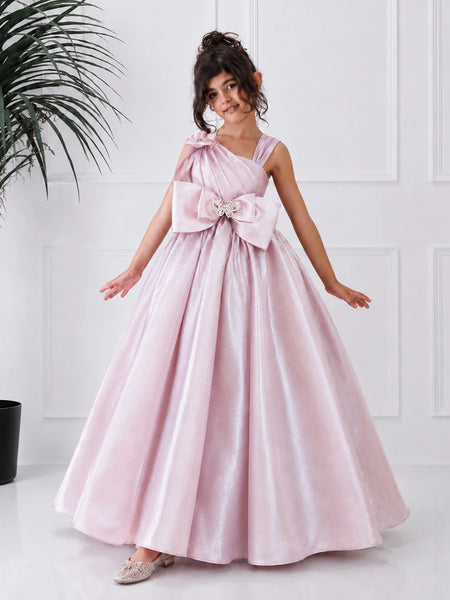 Young girl in a pink formal gown with a large bow standing against a white wall.