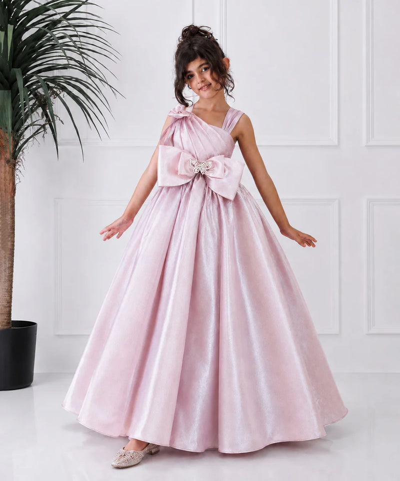 Young girl in a pink formal gown with a large bow standing against a white wall.