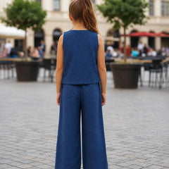 A young girl viewed from behind wearing a slate blue sleeveless top and matching wide-leg trousers, standing in a paved outdoor plaza with cafe tables and trees in the background.