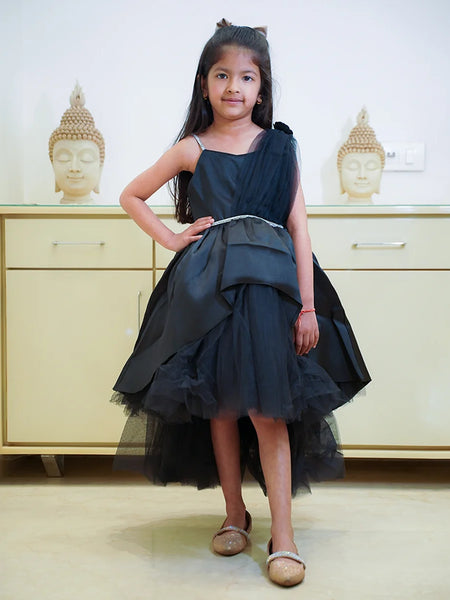 A young girl wearing a black party wear frock with a bow and pleated design, standing in front of a cabinet with Buddha statues on top.