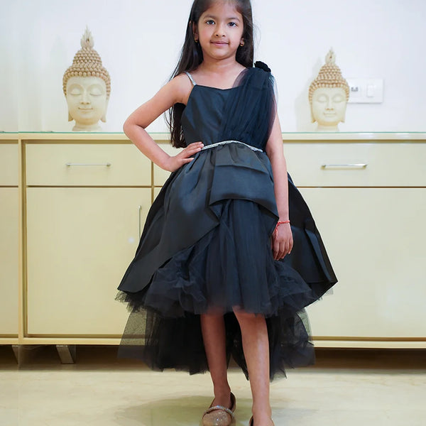 A young girl wearing a black party wear frock with a bow and pleated design, standing in front of a cabinet with Buddha statues on top.