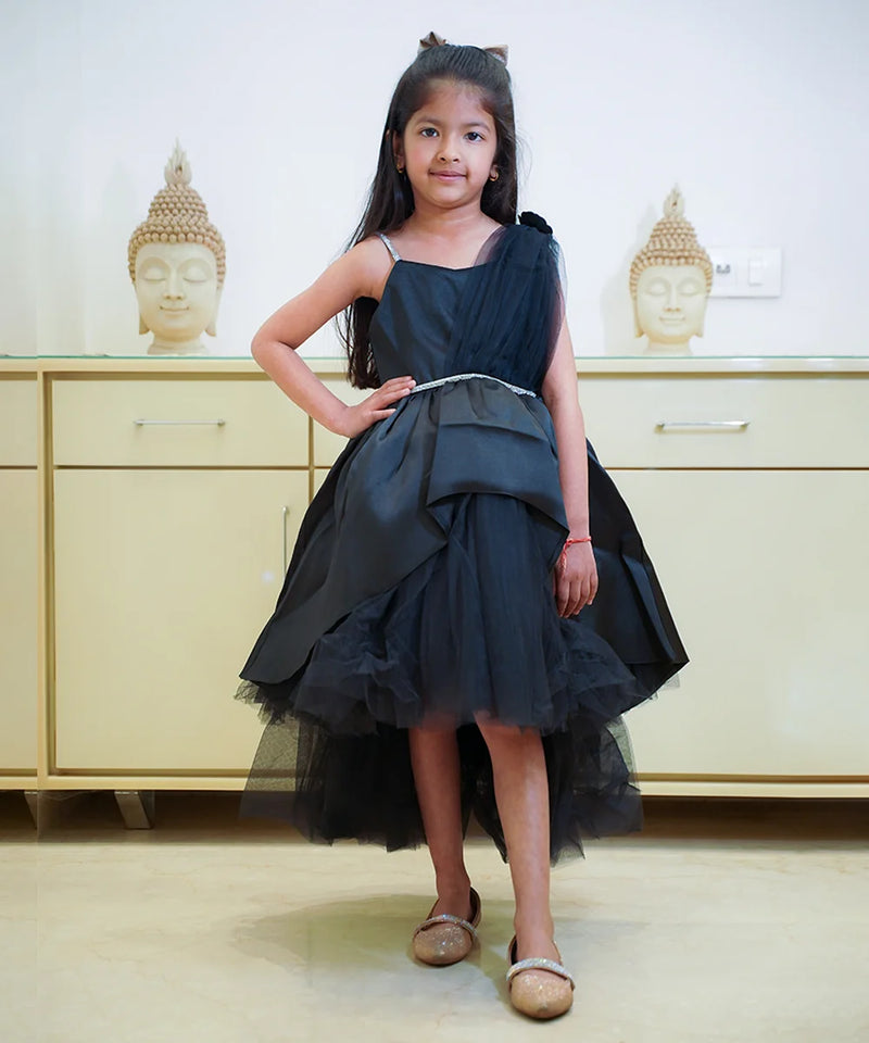 A young girl wearing a black party wear frock with a bow and pleated design, standing in front of a cabinet with Buddha statues on top.