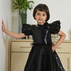 Young girl wearing a black dress with puffed sleeves and feather details, standing indoors.