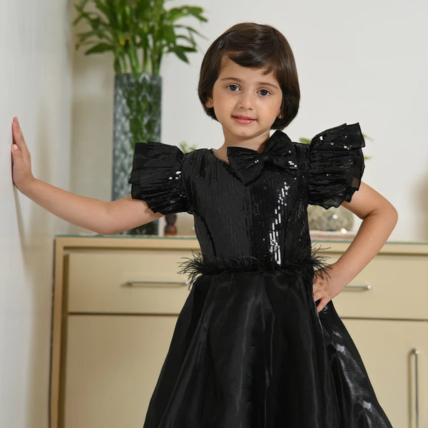 Young girl wearing a black dress with puffed sleeves and feather details, standing indoors.