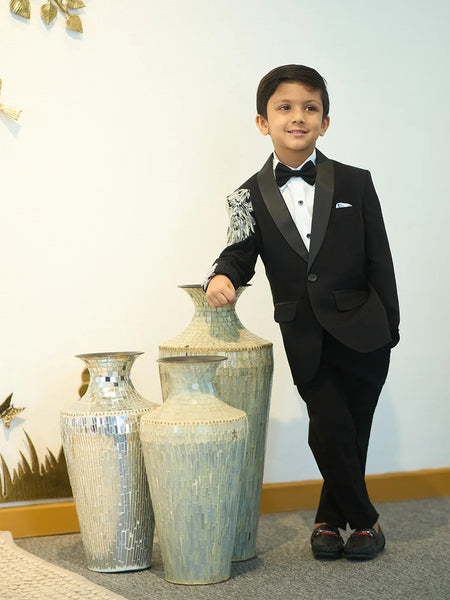 A boy in a black tuxedo with embroidered detailing on the sleeves, standing next to large vases.