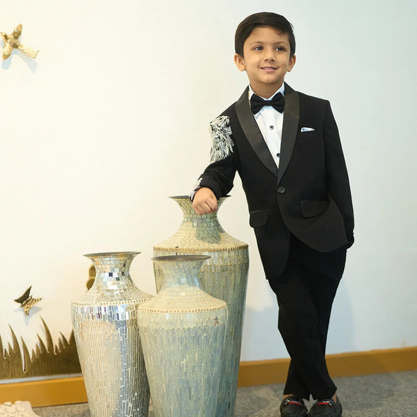 A boy in a black tuxedo with embroidered detailing on the sleeves, standing next to large vases.