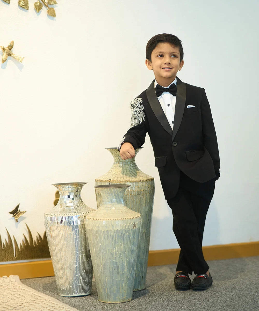 A boy in a black tuxedo with embroidered detailing on the sleeves, standing next to large vases.