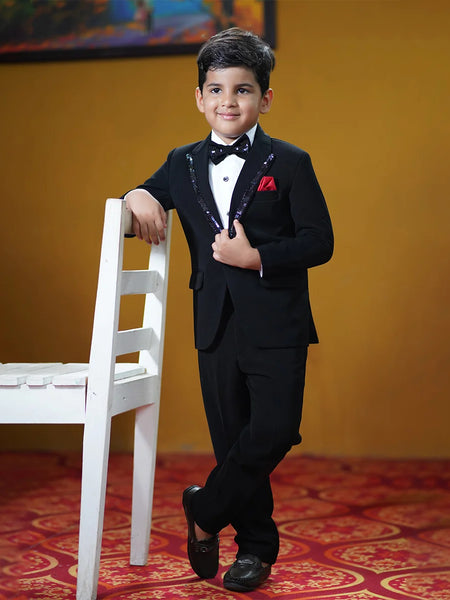 This classic tuxedo is rendered in black color and comes with matching pants, a white shirt, a black bow and a red pocket square. It features a beautiful sequin detailing on the collar and a bow curated from a black sequin fabric.