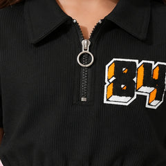A young girl with dark hair wearing a black short-sleeve crop top with a collar, a front zipper, and a graphic '84' on the chest, paired with blue jeans, posing against a pink background with decorative flower elements.