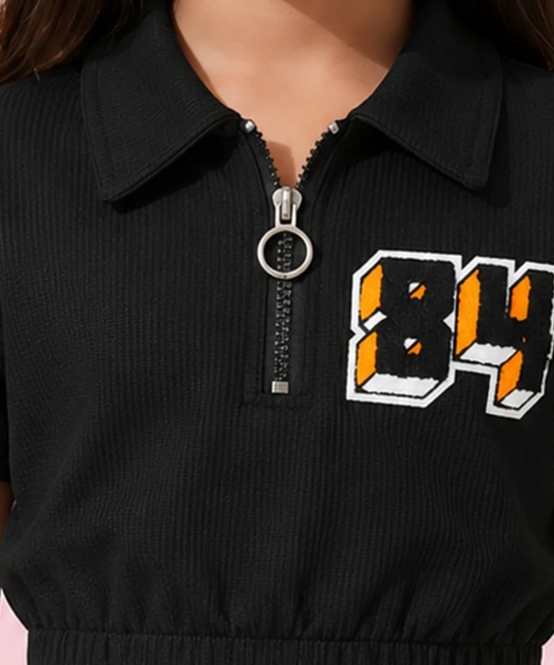 A young girl with dark hair wearing a black short-sleeve crop top with a collar, a front zipper, and a graphic '84' on the chest, paired with blue jeans, posing against a pink background with decorative flower elements.