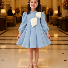 A young girl stands in a grand hotel lobby wearing a light blue dress with a large cream bow on the chest, paired with gold shoes.