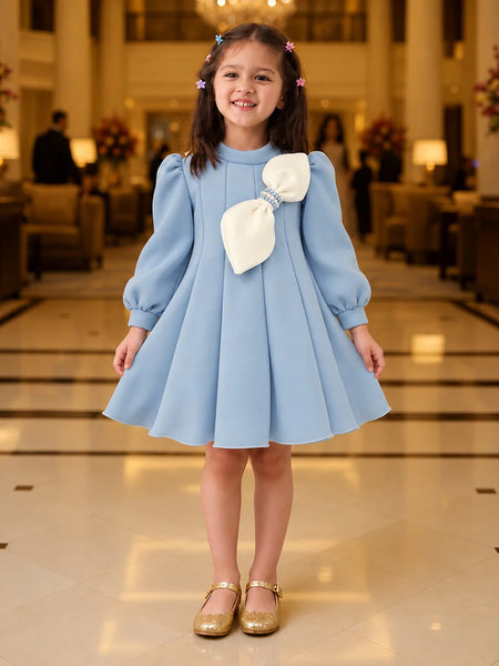 A young girl stands in a grand hotel lobby wearing a light blue dress with a large cream bow on the chest, paired with gold shoes.