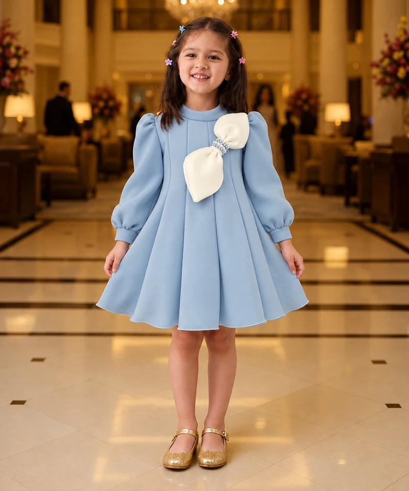 A young girl stands in a grand hotel lobby wearing a light blue dress with a large cream bow on the chest, paired with gold shoes.