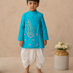 A young boy stands barefoot wearing a bright turquoise traditional outfit with white dhoti-style pants and gold trim, posed against a warm beige studio backdrop next to a ceramic vase with white flowers.