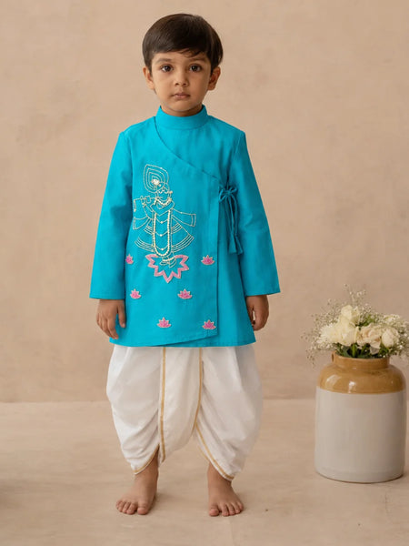 A young boy stands barefoot wearing a bright turquoise traditional outfit with white dhoti-style pants and gold trim, posed against a warm beige studio backdrop next to a ceramic vase with white flowers.
