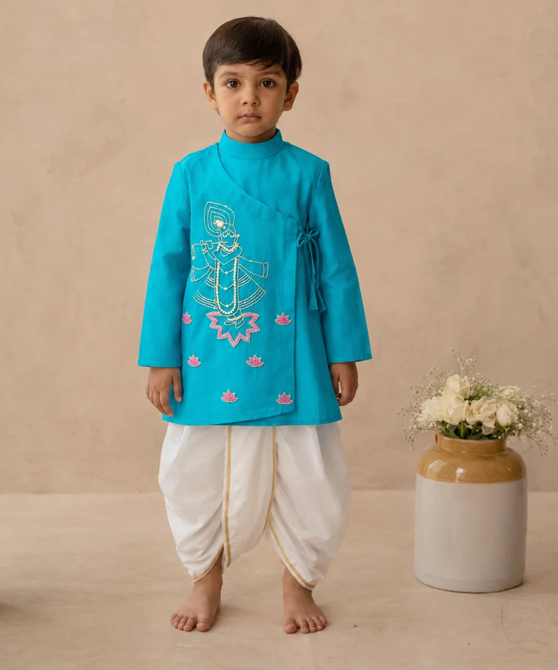 A young boy stands barefoot wearing a bright turquoise traditional outfit with white dhoti-style pants and gold trim, posed against a warm beige studio backdrop next to a ceramic vase with white flowers.
