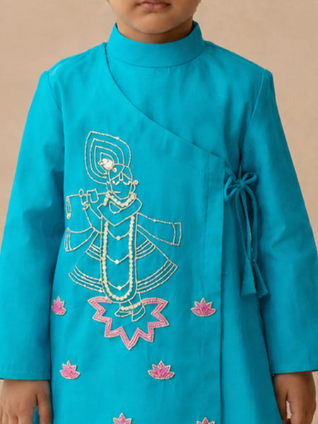 A child wearing a bright turquoise blue wrap-style kurta with white and pink embroidered details depicting a traditional figure and lotus flowers, set against a plain beige background.