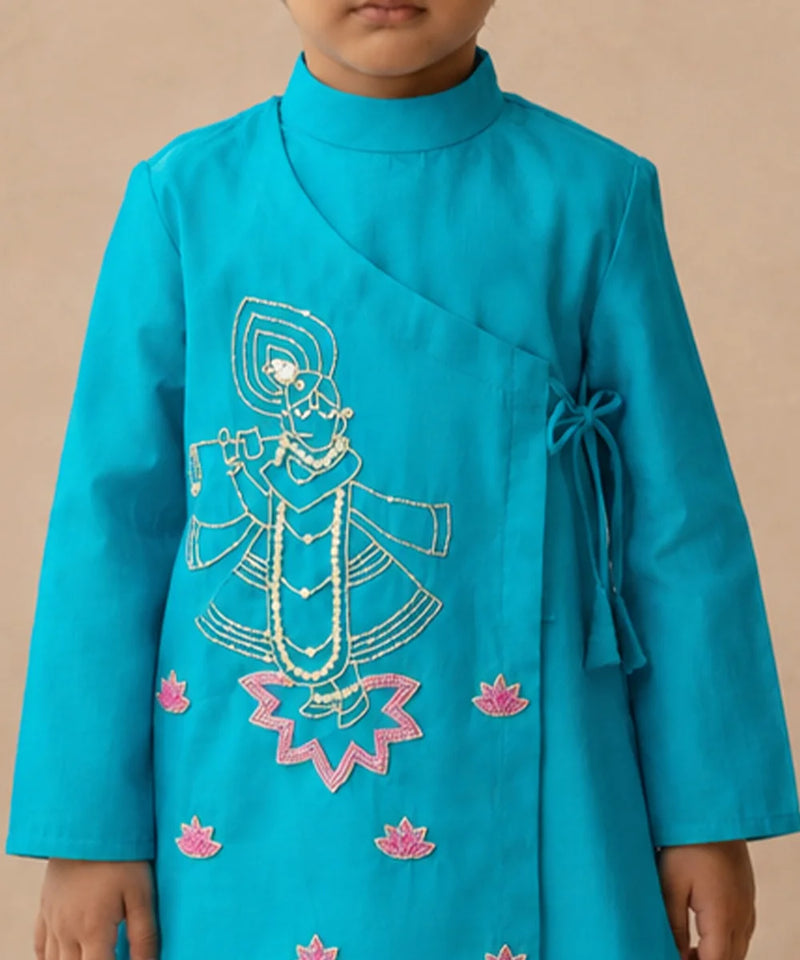 A child wearing a bright turquoise blue wrap-style kurta with white and pink embroidered details depicting a traditional figure and lotus flowers, set against a plain beige background.