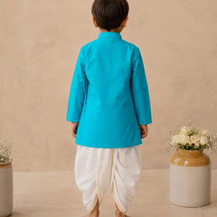 A child viewed from the back wearing a bright turquoise-blue kurta and white dhoti pants, standing barefoot against a warm beige textured wall with decorative ceramic vases on either side.